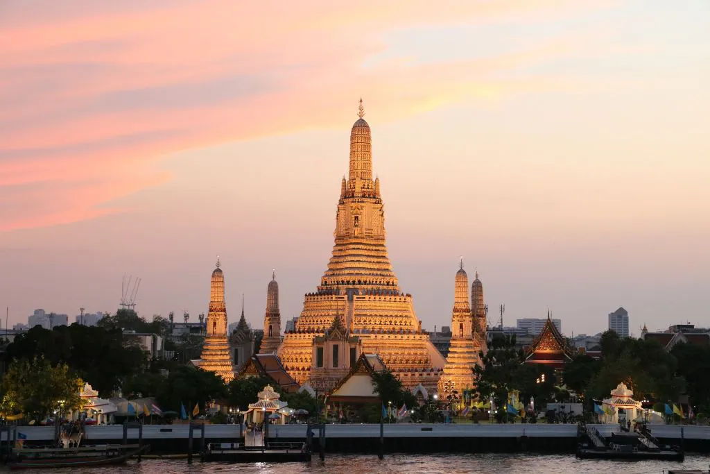 Temple Wat Arun à Bangkok illuminé au coucher du soleil, avec un ciel rose orangé en arrière-plan.